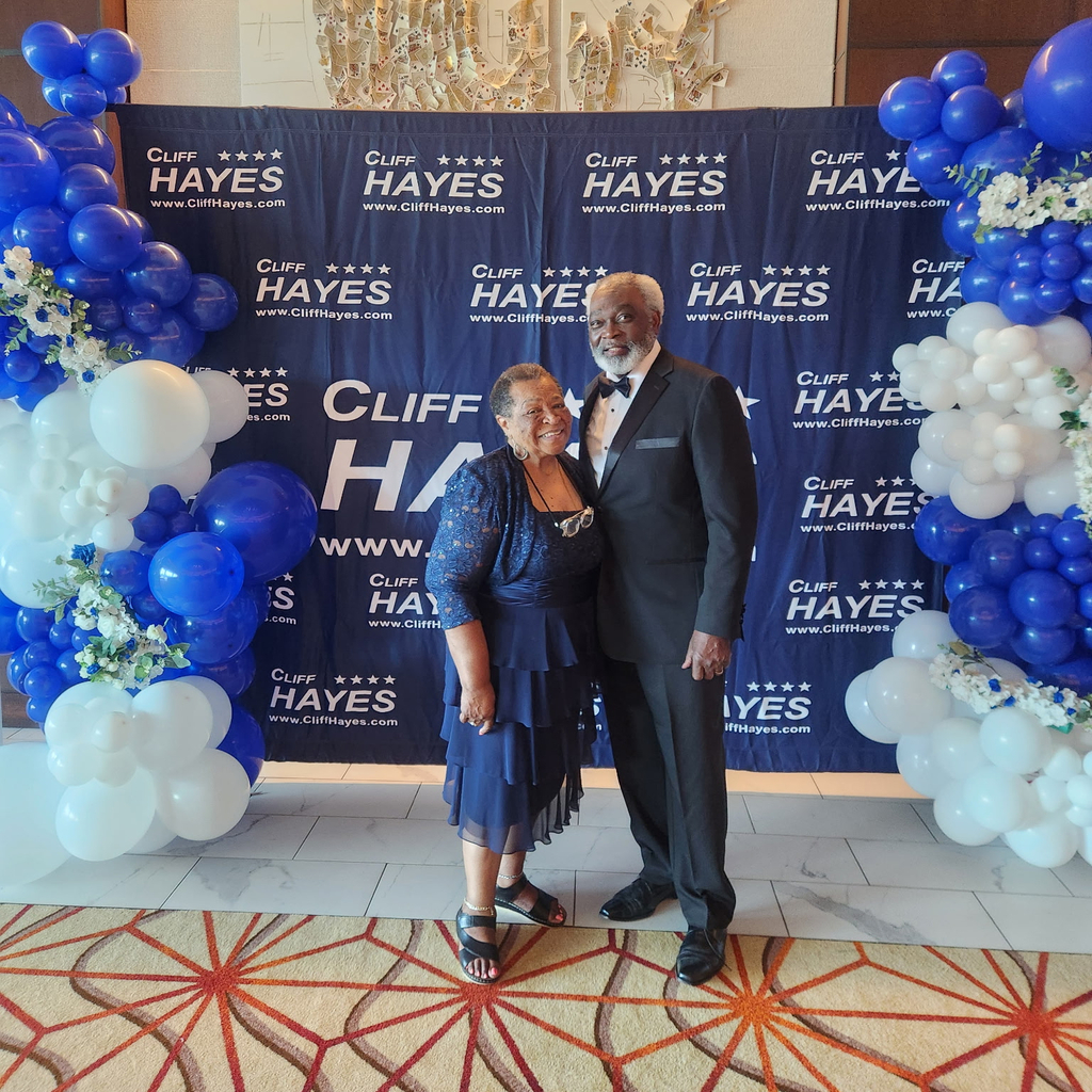 A smiling, elegantly dressed older Black couple standing in front of a dark blue backdrop repeated with "Cliff Hayes" and four stars. The woman wears a sleeveless dark blue layered lace dress, and the man wears a classic dark suit with a black bow tie. They are framed on both sides by large blue, white, and floral balloon arches.