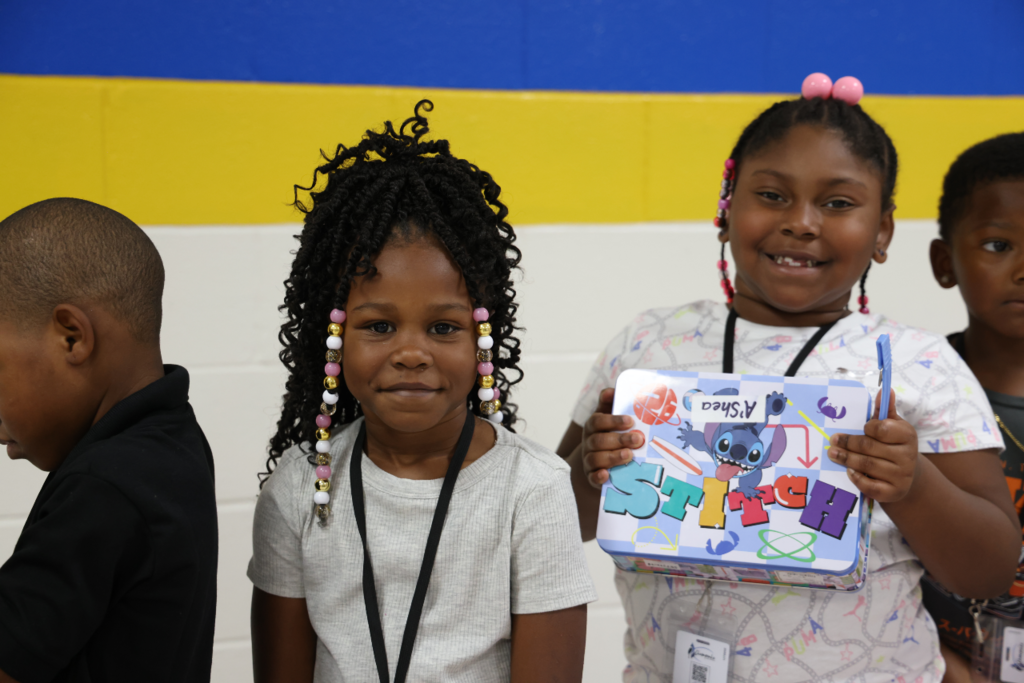 A young student proudly holding up a Stitch lunch box or tin next to a classmate.
