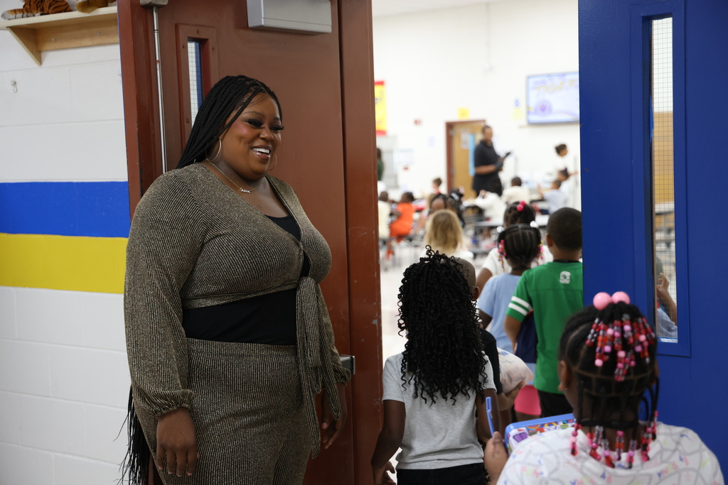 A smiling staff member in a glittery gold top standing near a doorway as a line of elementary students walks into a classroom.
