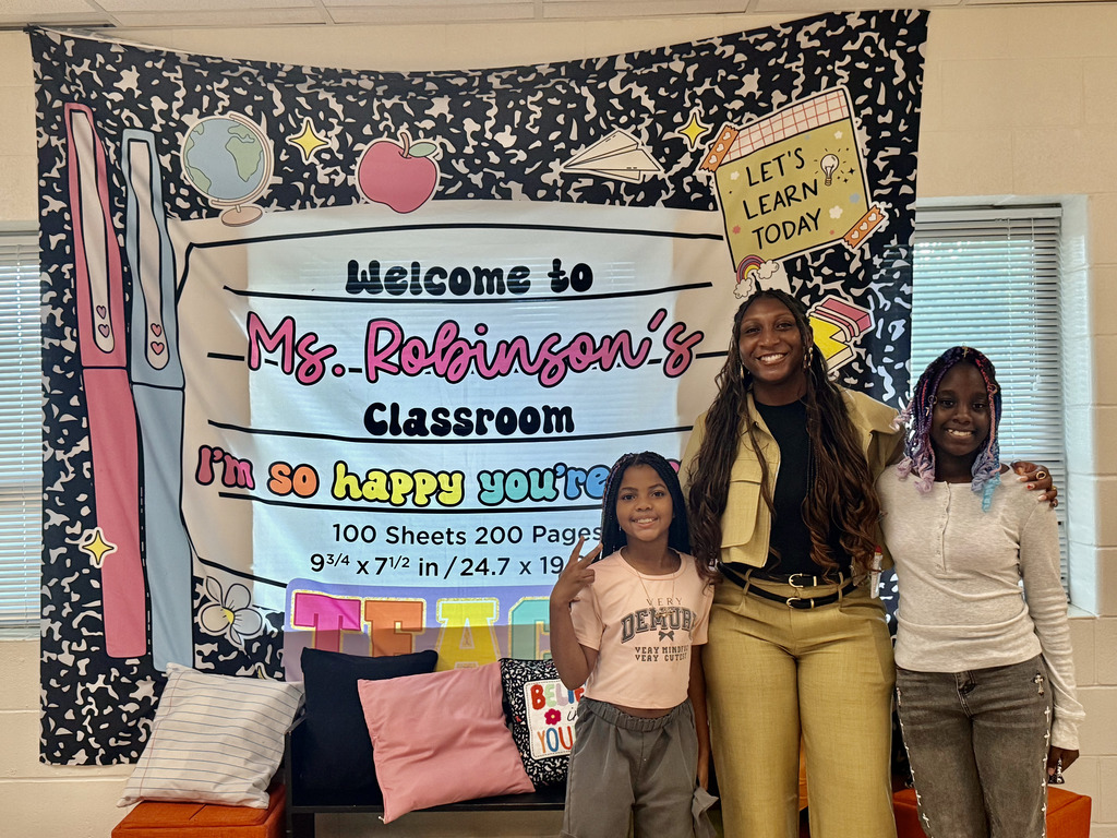 A teacher and two young students smiling together in front of a classroom banner styled like a composition notebook that reads "Welcome to Ms. Robinson's Classroom."