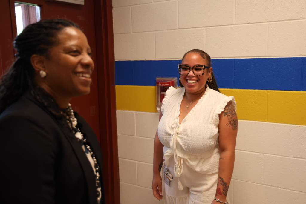 Two women laughing together in a school hallway featuring blue and yellow wall stripes.