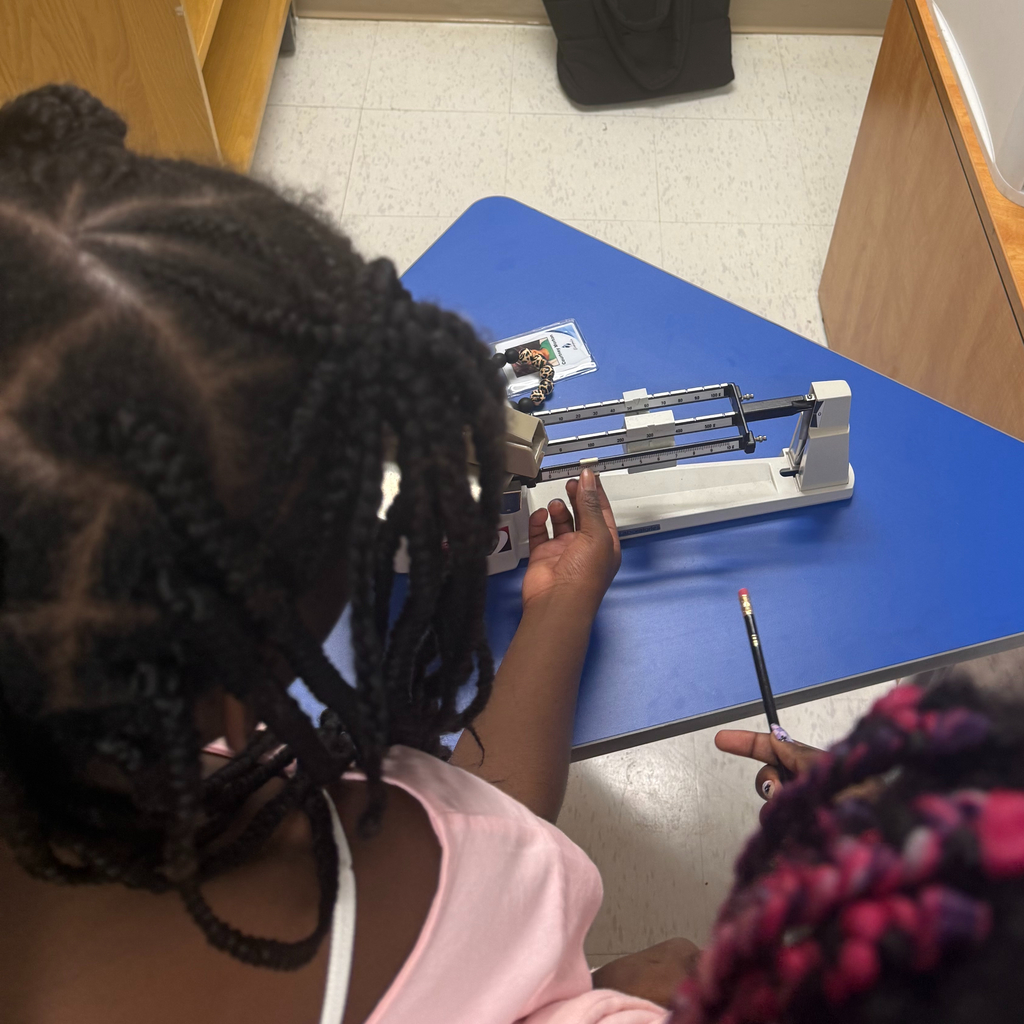 Over-the-shoulder view of a student adjusting a triple beam balance on a blue desk during a science activity.