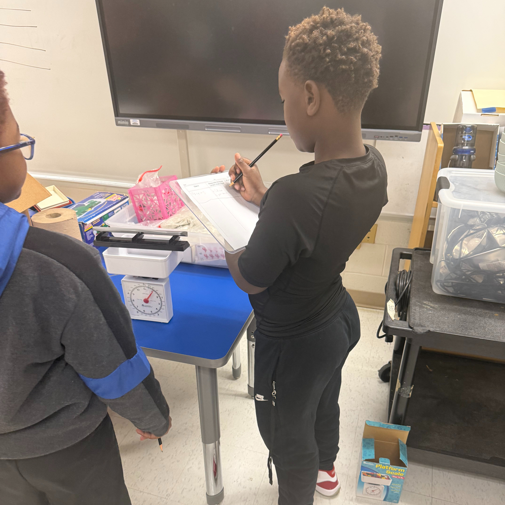 A young student writes on a clip-board worksheet next to a small tabletop scale in a classroom, with a classmate watching nearby.