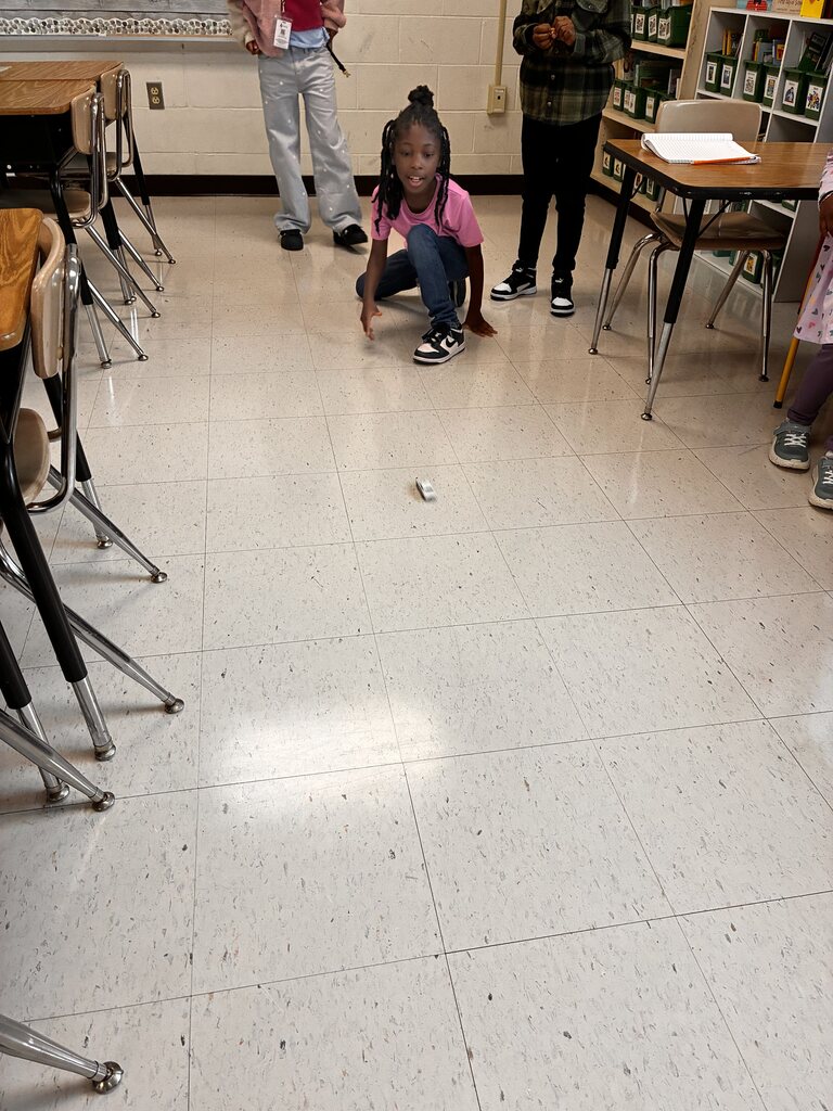A student crouches on a classroom floor, watching a small toy car roll across the open tile space.