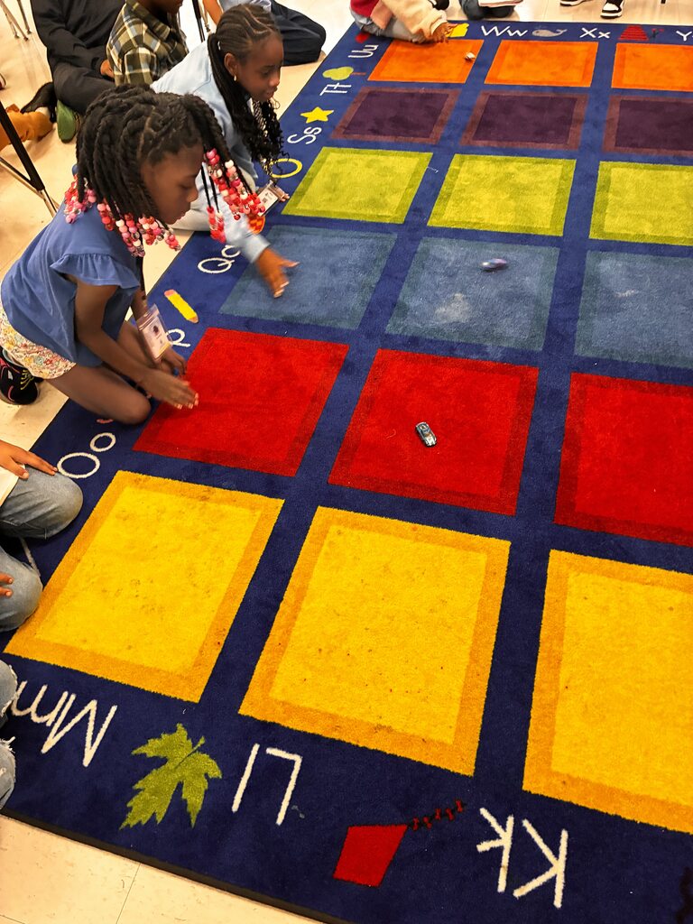 High-angle shot of young students sitting on the floor around a colorful classroom carpet patterned with brightly colored squares and alphabet letters. One child reaches out as a small toy car rolls across the center squares.