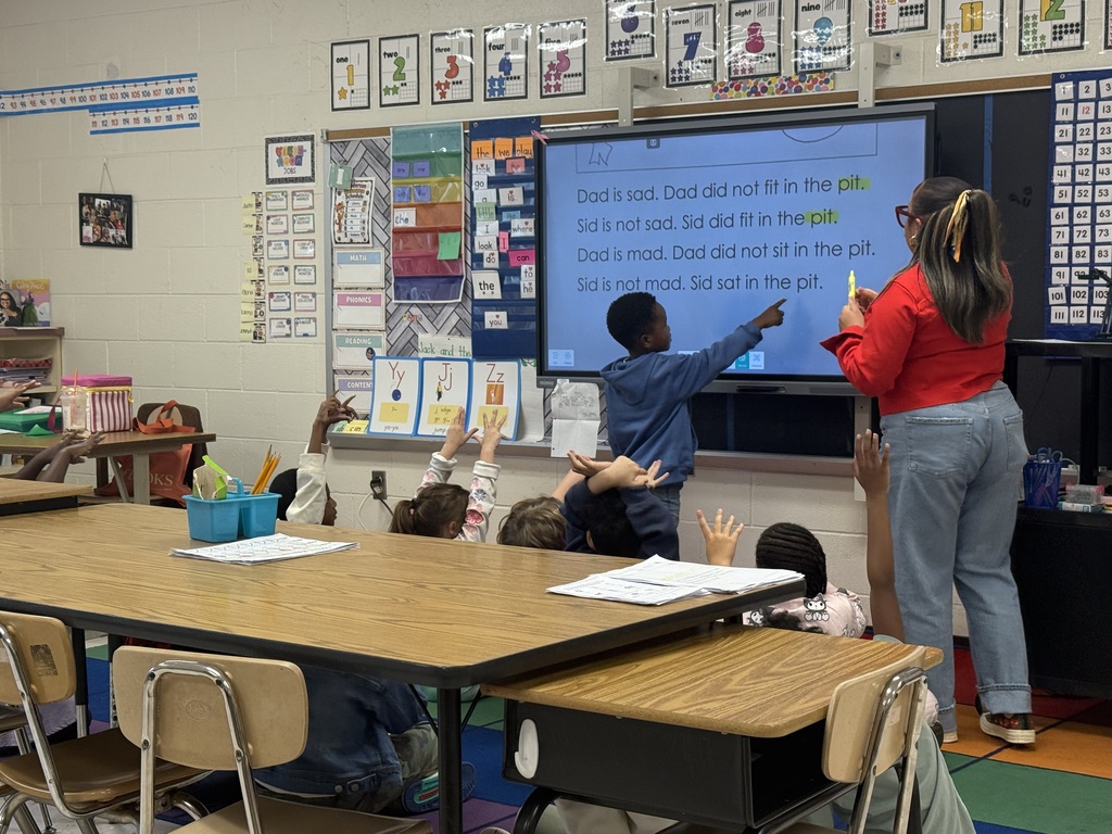 An elementary school classroom during a phonics lesson. A young boy stands at an interactive smartboard pointing to a short story about "Dad and Sid in the pit," while a female teacher in a red sweater and jeans stands beside him with a yellow marker. Surrounding students sit at tables with their hands raised to answer.