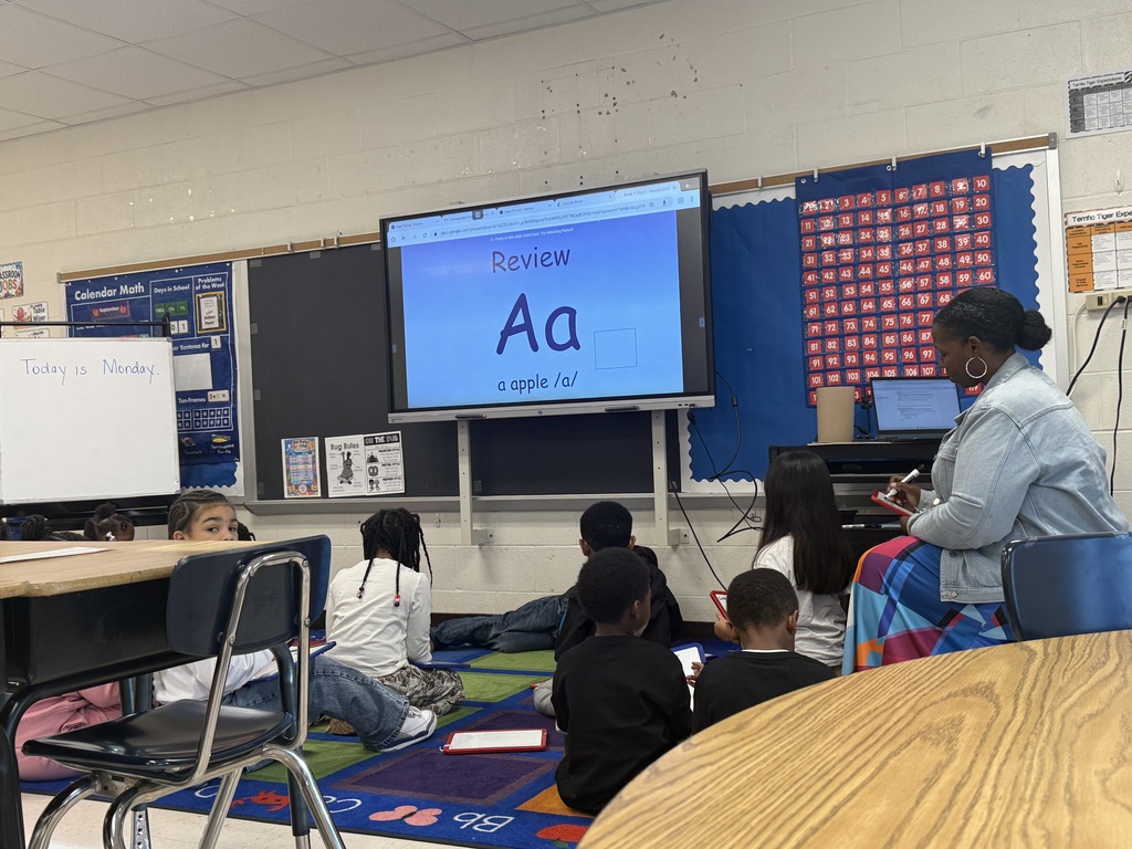 A female teacher sitting on a stool in front of an interactive display screen showing a letter review slide for the letter "Aa" with the prompt "a apple /a/". Several young students sit on a colorful classroom rug facing the screen and listening attentively.