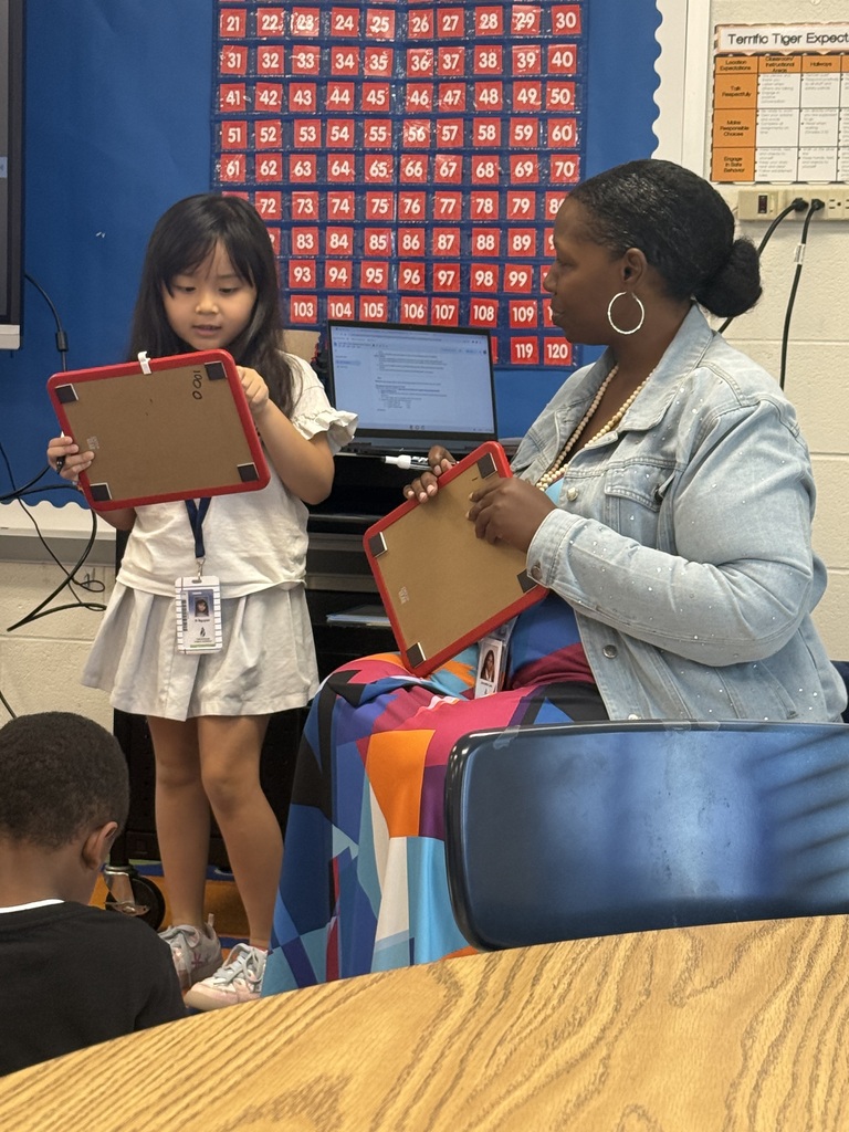 A teacher in a denim jacket and colorful skirt sits next to a young female student. Both are holding small dry-erase whiteboards framed in red plastic and comparing their written work in a bright elementary classroom setting.