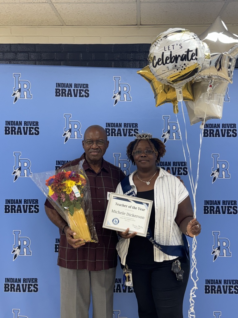 Michele Dickerson, Indian River Middle School’s Teacher of the Year, with her father, smiles while holding her Teacher of the Year certificate and gold and white balloons. She wears a “Teacher of the Year” sash and tiara while standing in front of an Indian River Braves backdrop.