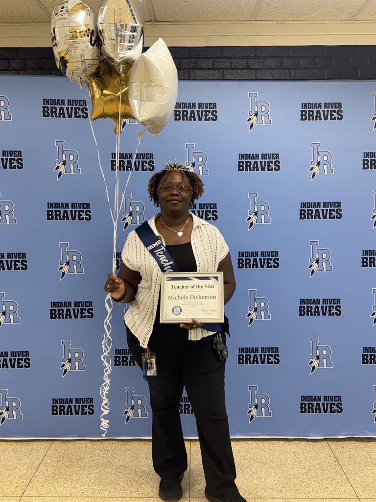 Michele Dickerson, Indian River Middle School’s Teacher of the Year, smiles while holding her Teacher of the Year certificate and gold and white balloons. She wears a “Teacher of the Year” sash and tiara while standing in front of an Indian River Braves backdrop.