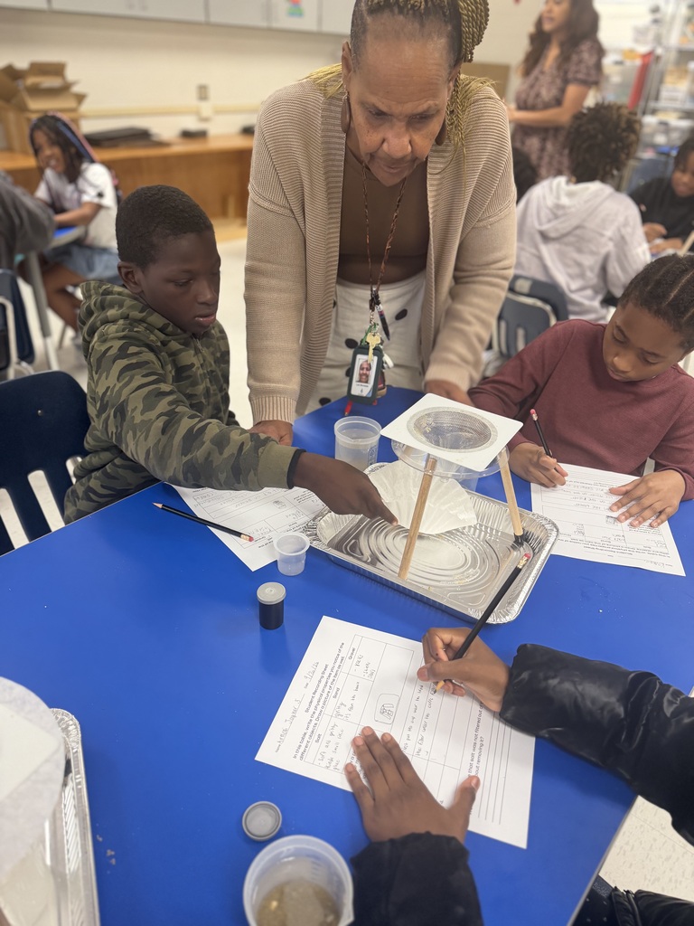 An adult standing over two students at a blue classroom table, guiding them through a science experiment with a homemade filtration setup made of coffee filters, plastic cups, and wooden craft sticks. The students hold pencils and work on accompanying lab worksheets.