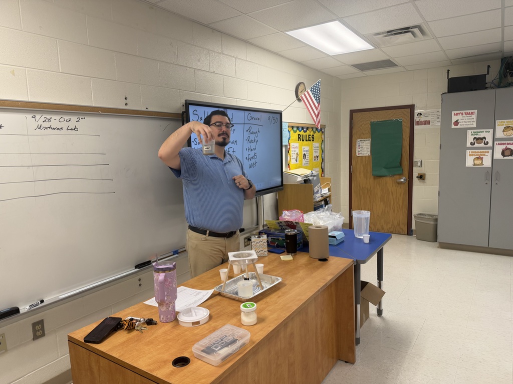 A teacher standing at the front of a classroom next to a wooden desk, holding up a clear plastic cup containing a liquid mixture to demonstrate a experiment to the class. On the whiteboard behind him, text reads "9/28-Oct 2" and "Mixtures Lab".