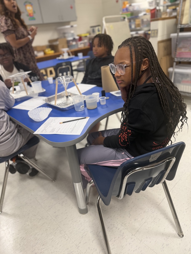 A student wearing clear-framed glasses and a black hoodie sitting at a blue classroom table during a science activity. A homemade funnel and filter setup sits on a tray on the table alongside plastic cups and lab papers.