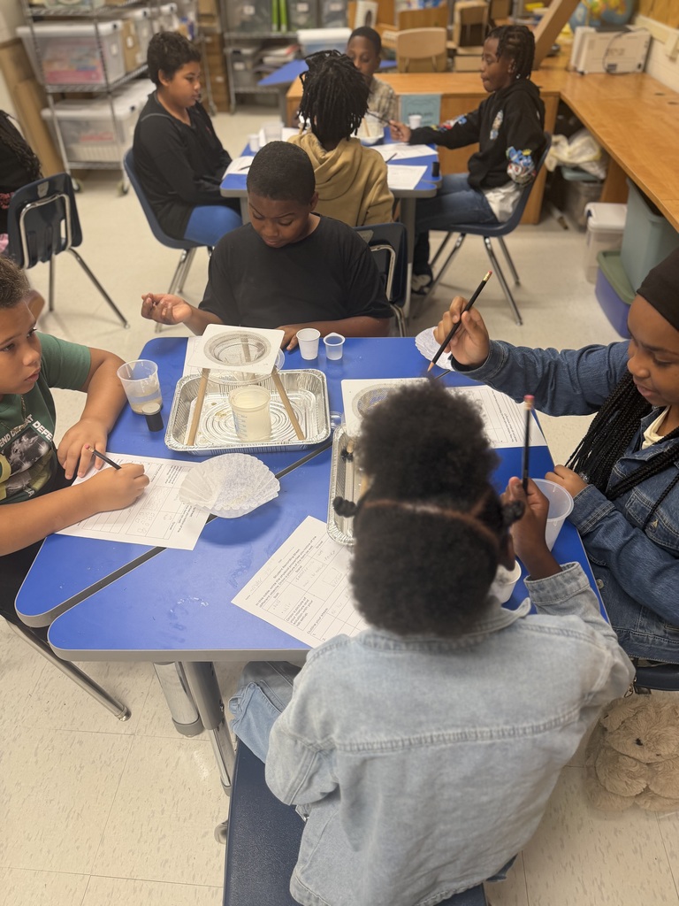 Four students seated around a blue table in a science classroom, using pencils to write on lab worksheets while observing a makeshift filtration setup placed in a foil tray at the center of the table
