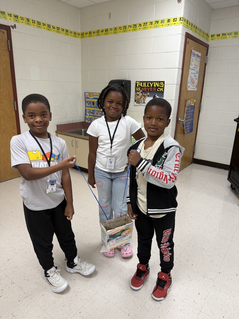 Three elementary students standing in a line in a classroom, presenting a small container decorated with stickers suspended between two long blue pipe cleaners or strings held by the students on either end.