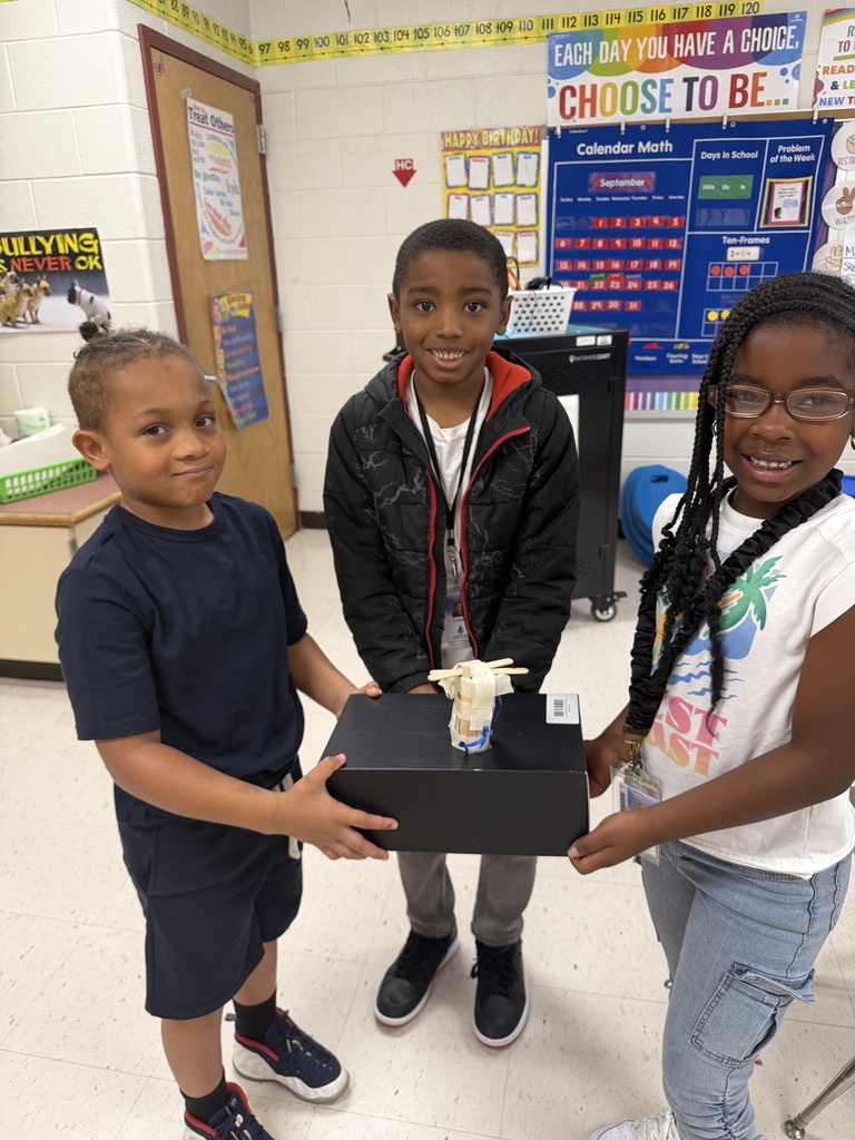 Three smiling elementary students displaying a STEM project together in a classroom. The students on the left and right support a black shoe box with a small wooden craft structure secured to the top, while the student in the center stands behind them.