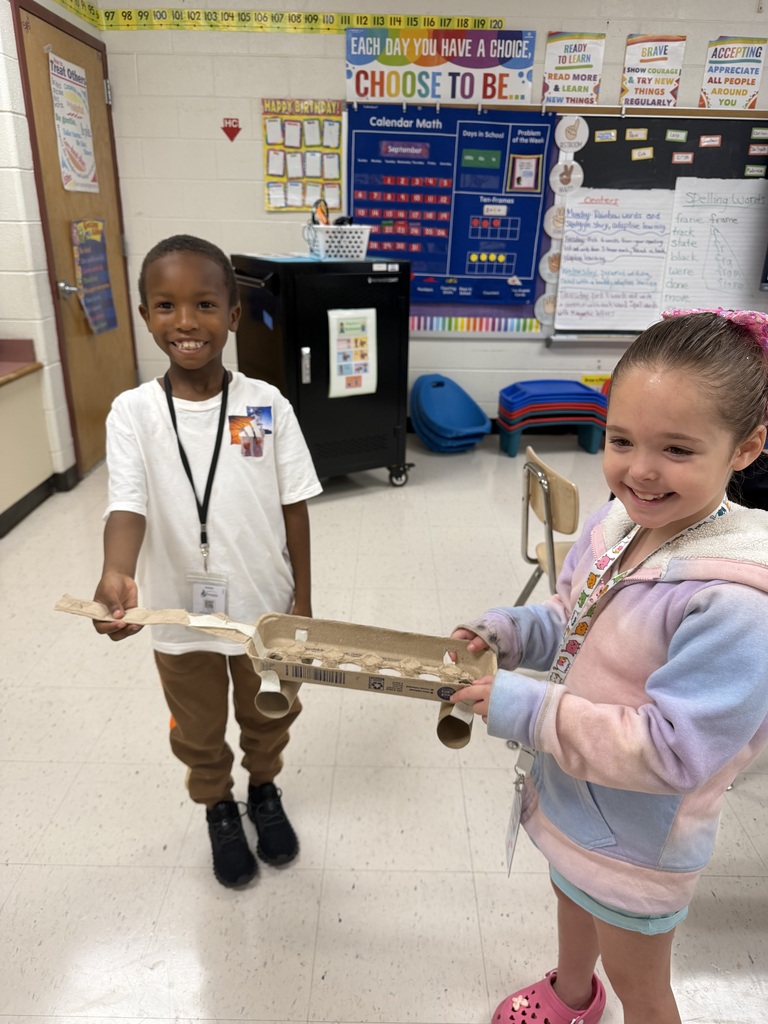 Two young elementary students smiling and standing together in a classroom, holding up a handmade craft project constructed from an egg carton, paper towel rolls, and tape. Educational charts decorated with calendar math and motivational posters line the classroom walls in the background.