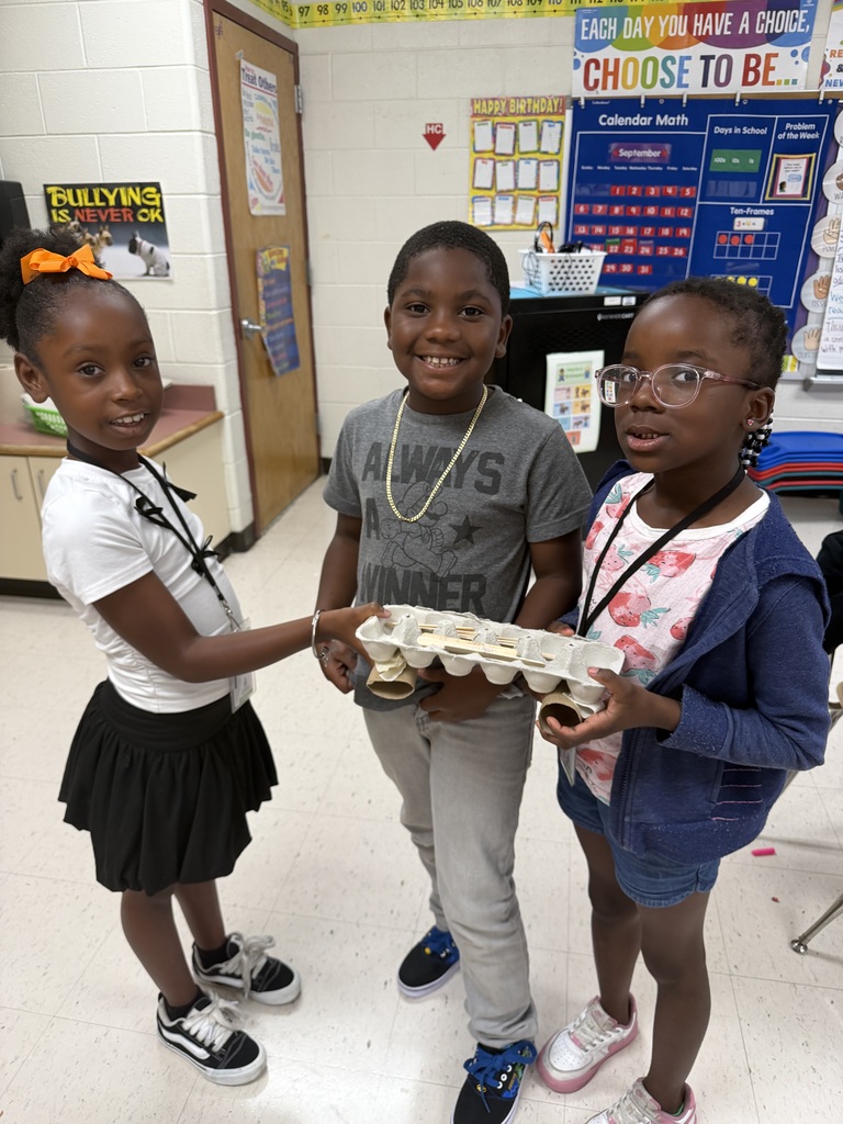 Three elementary students standing together in a classroom and proudly holding a model bridge or vehicle project crafted from an egg carton, cardboard tubes, and popsicle sticks.