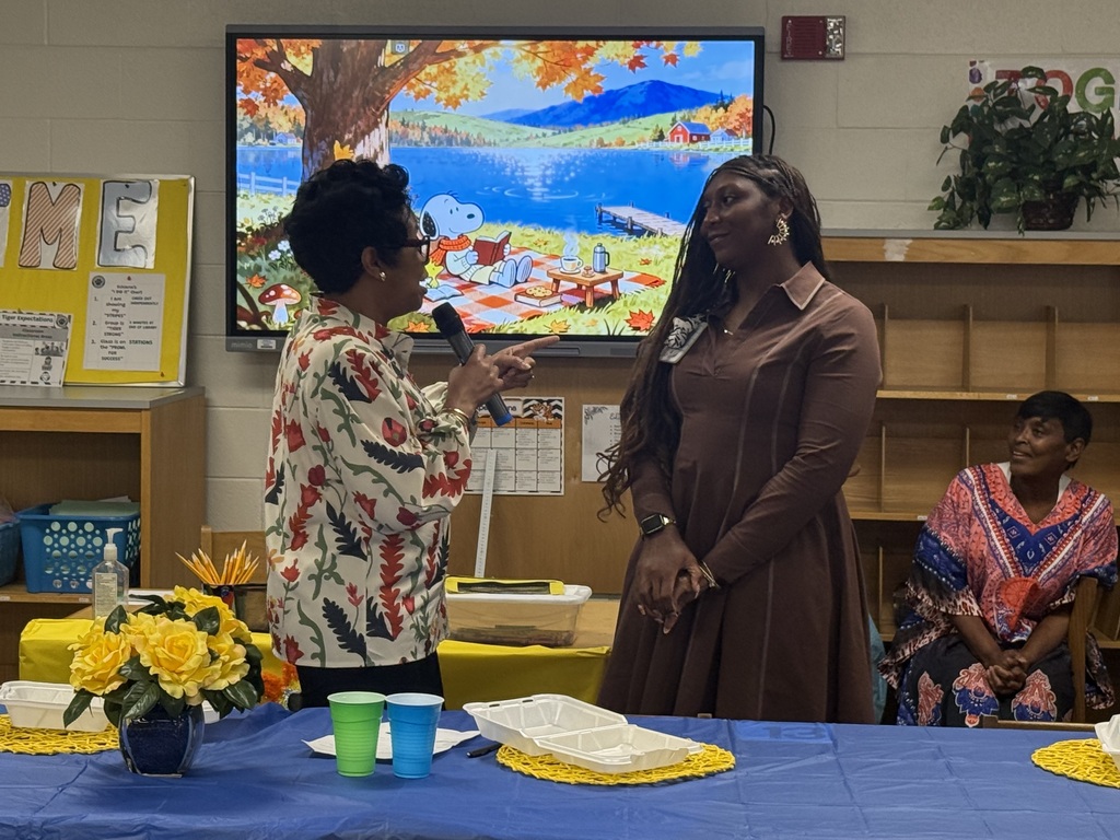 A speaker holding a microphone addresses Ms. Aubreya Robinson during a indoor celebration, with a digital display screen featuring a autumn cartoon illustration in the background.