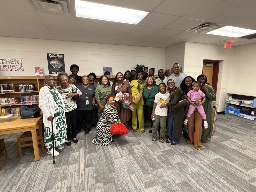 Large group photo of school staff, colleagues, and family gathered inside a school library to celebrate Ms. Aubreya Robinson, who stands in the center holding gifts and flowers.