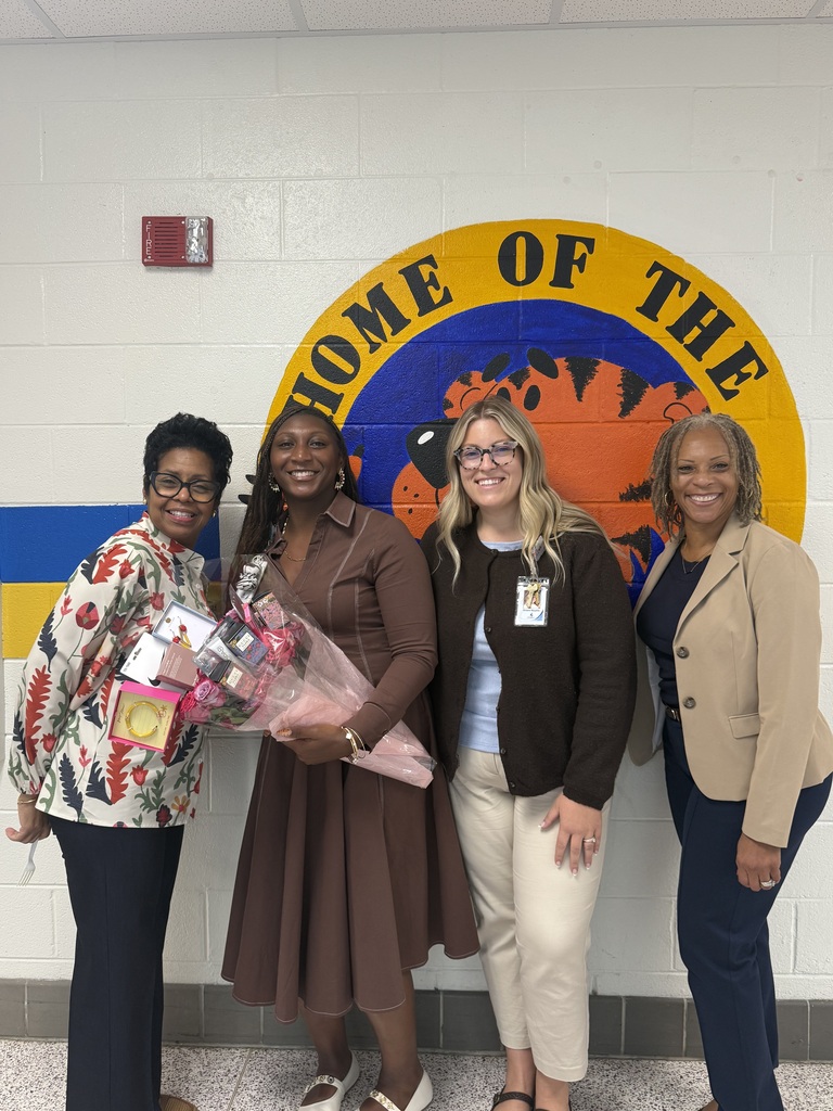 Group photo of Ms. Aubreya Robinson smiling alongside three colleagues in a school hallway. Ms. Robinson stands near the center holding flowers and gifts in front of a colorful school wall painting.