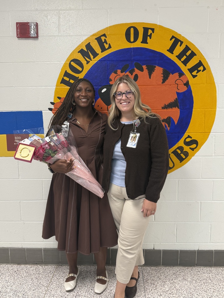 Ms. Aubreya Robinson standing in a school hallway with a smiling colleague. Ms. Robinson holds a bouquet of flowers and small gift items in front of a painted wall logo that reads "Home of the Cubs".