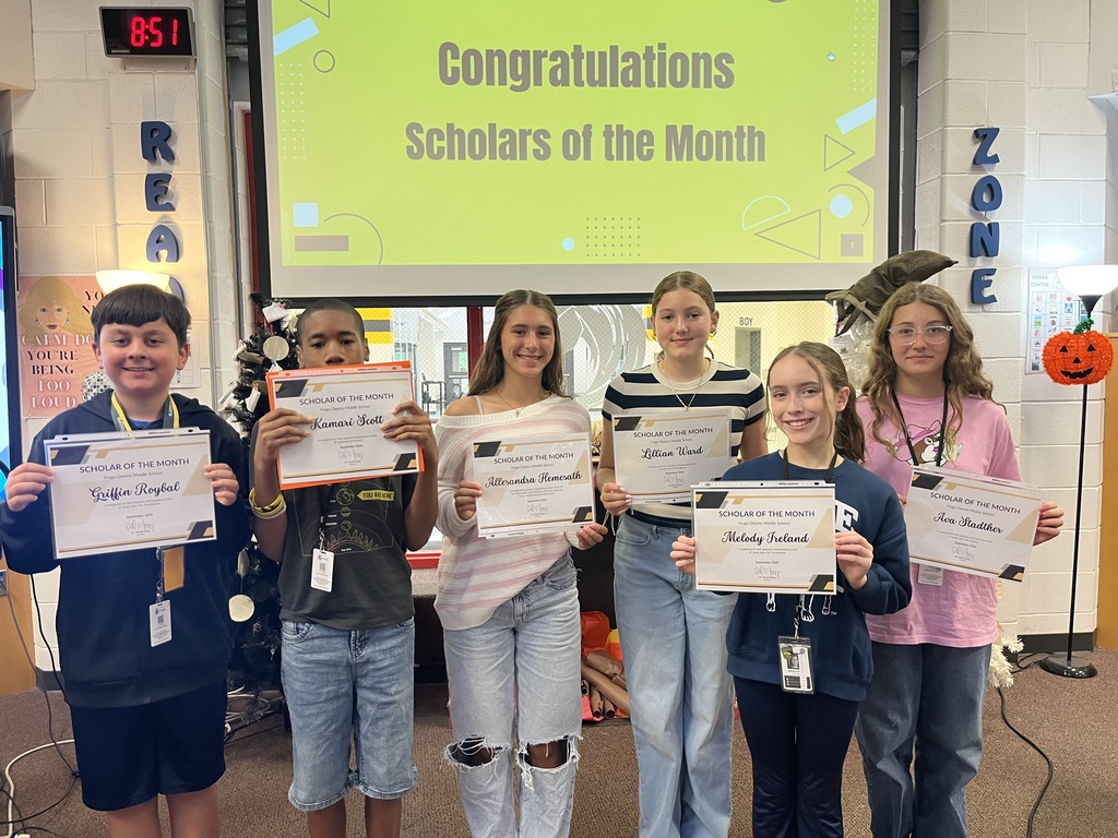 A group of six students standing in front of a board congratulating them on being scholars for the month of September.  Each student is holding up a certificate