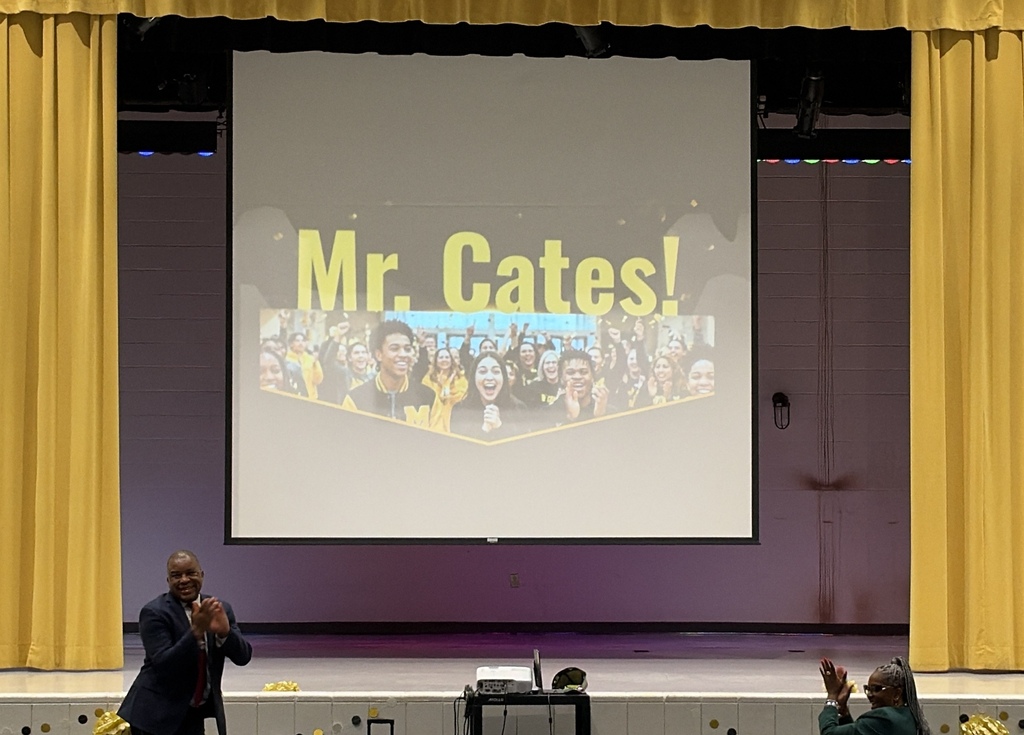 A photo of a screen with students and the name Mr. Cates to announce him as teacher of the year.  Another man in a suit is in the image.