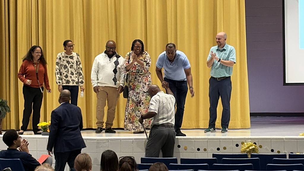 A picture of 6 adults on a stage congratulating a man not on the stage and another man in a black suit.