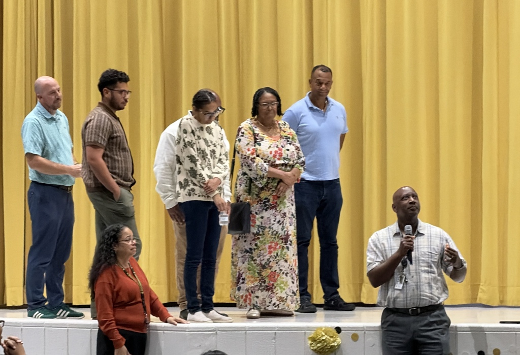 A picture of 6 adults on a stage listening to a man in a plaid white shirt that just won an award.
