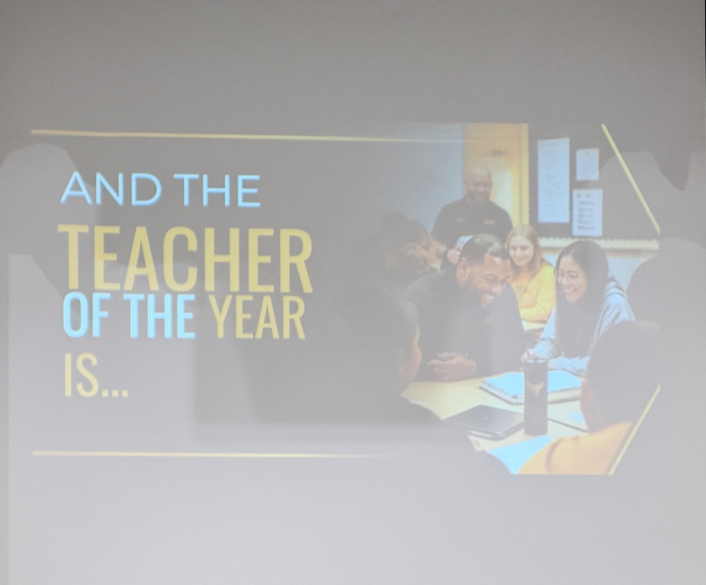 A photo of a screen with a teacher instructing students with the words "and the teacher of the year is..."