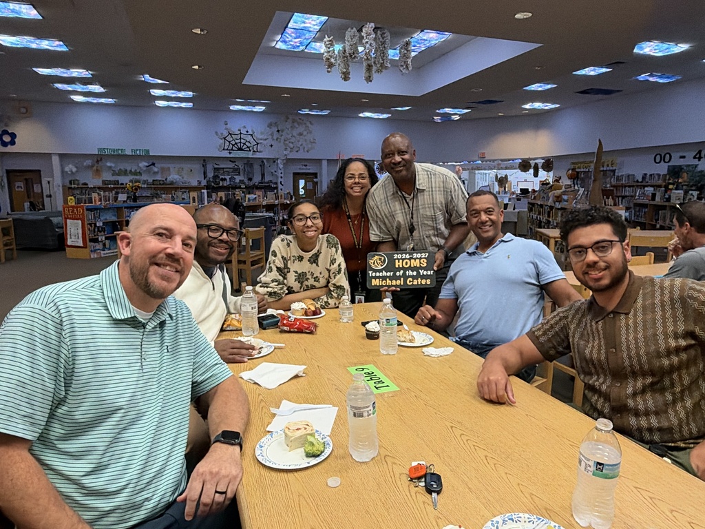 A group of seven adults sitting at a table with water bottles and plates of food.  One man is holding a sign that says teacher of the year.