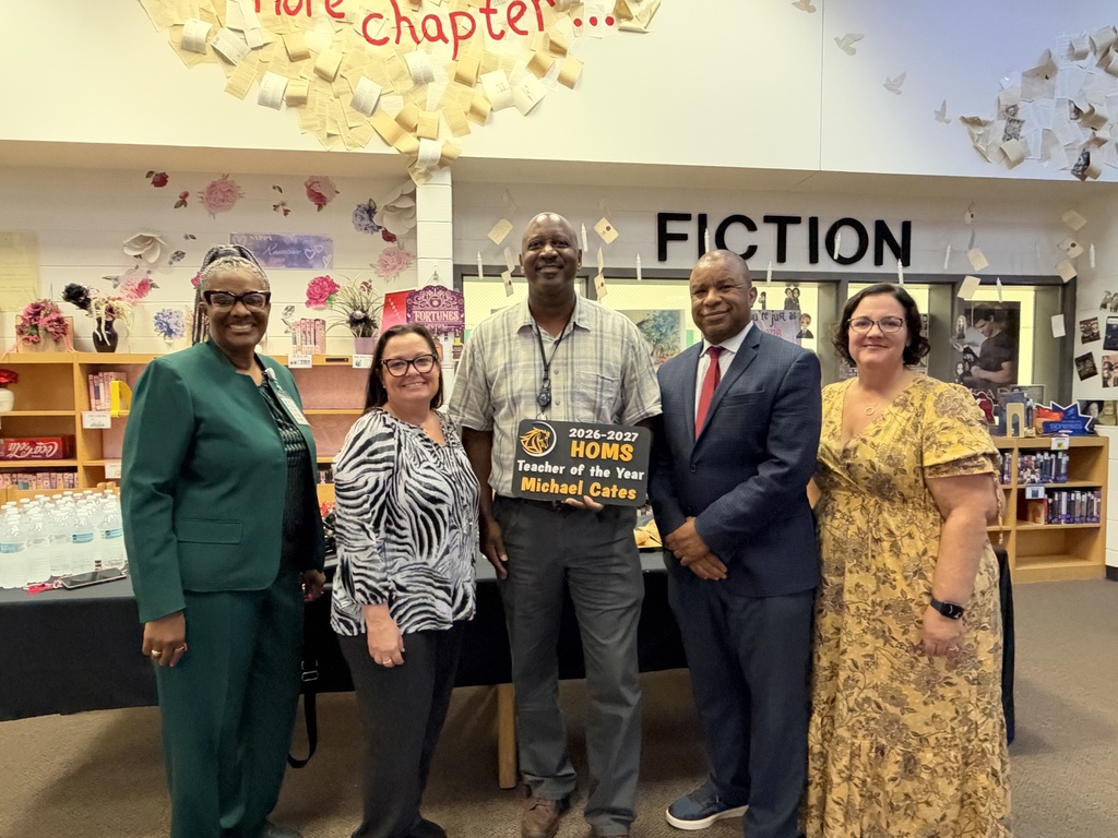 Five adults dressed in business-like attire.  The man in the middle is holding a sign that says teacher of the year.