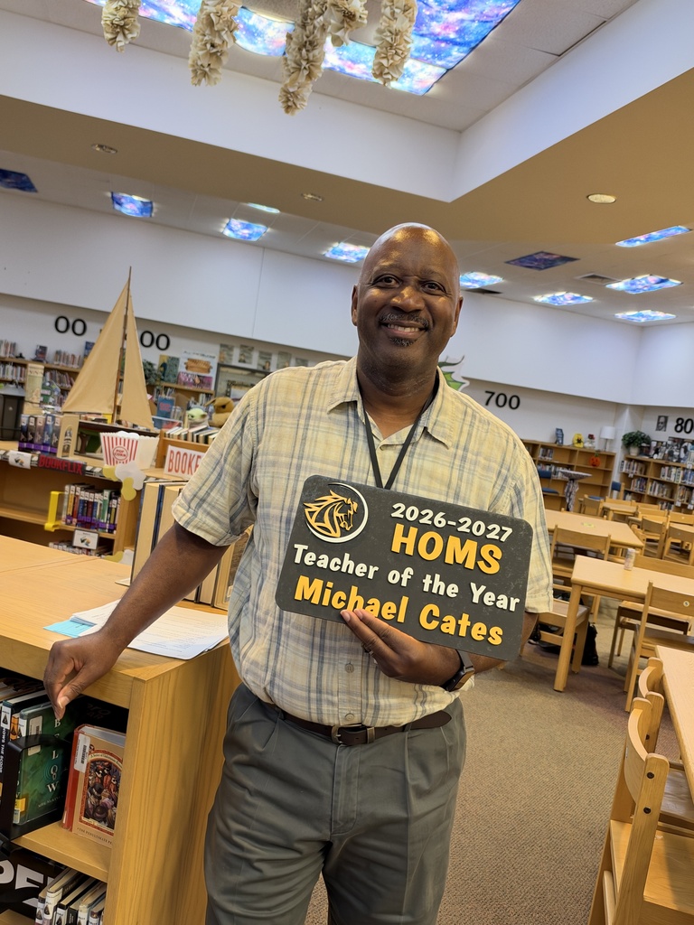 A man standing in a library dressed in a white plaid shirt, gray pants, holding a sign that says teacher of the year.
