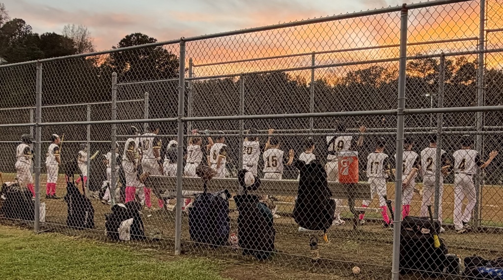 A baseball team wearing white uniforms with black numbers stands along a fence line watching a teammate up at bat.