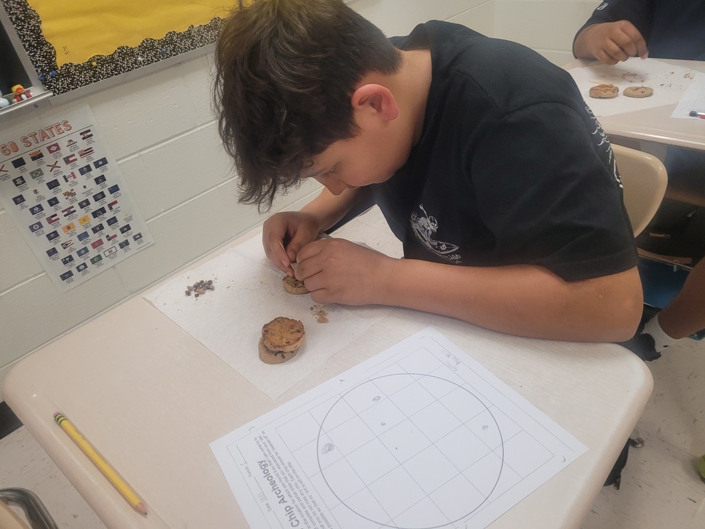 A student in a black shirt has his head down facing the desk breaking apart a cookie for an archaeology activity.