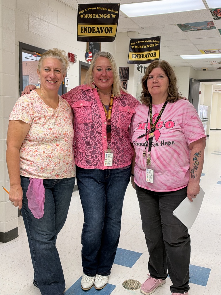 Three adult females standing in a hallway.  They are wearing pink shirts and jeans.