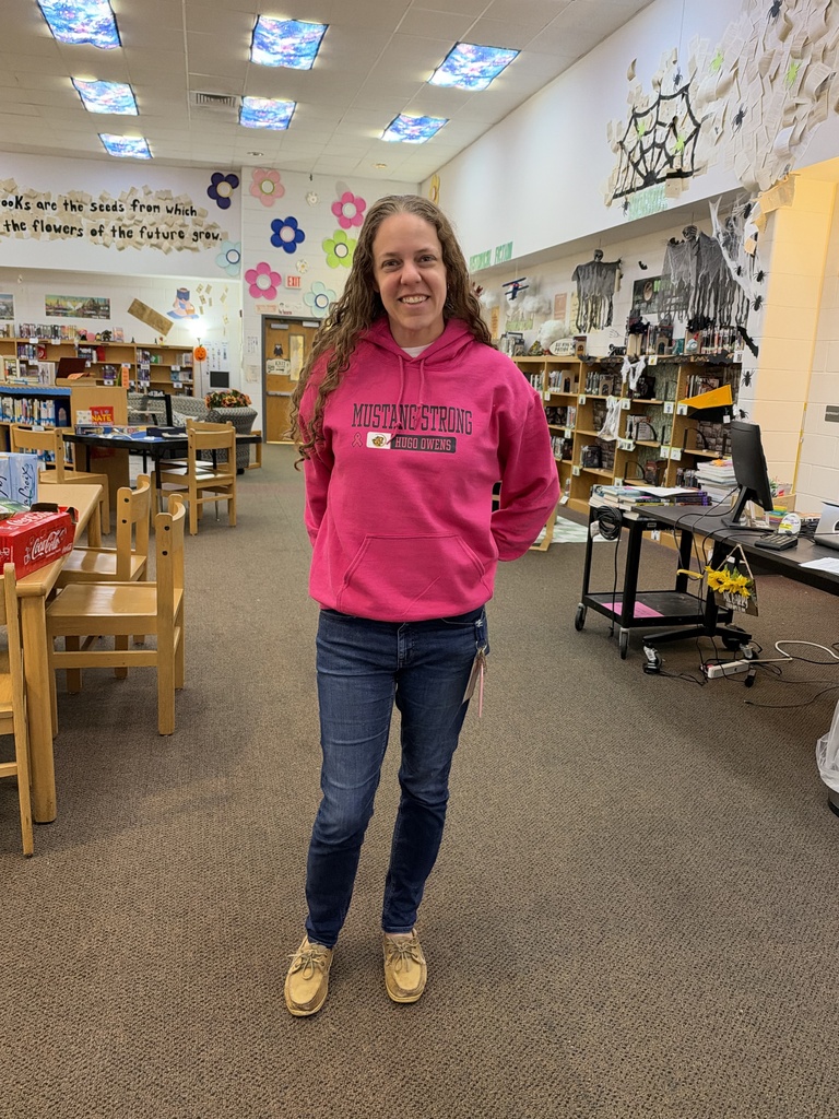 A worman standing in a library next to a desk wearing a bright pink sweatshirt and jeans.