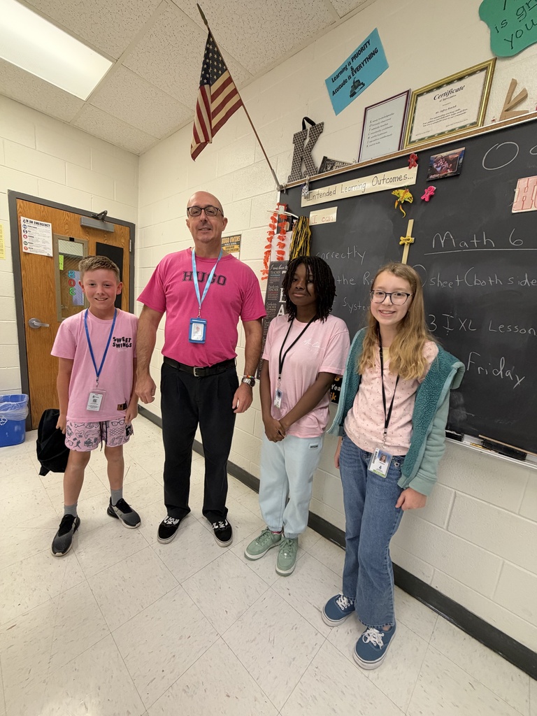 Three students and an adult male teacher standing next to a chalkboard.  The students are wearing light pink shirts and the teacher is wearing a bright pink shirt.