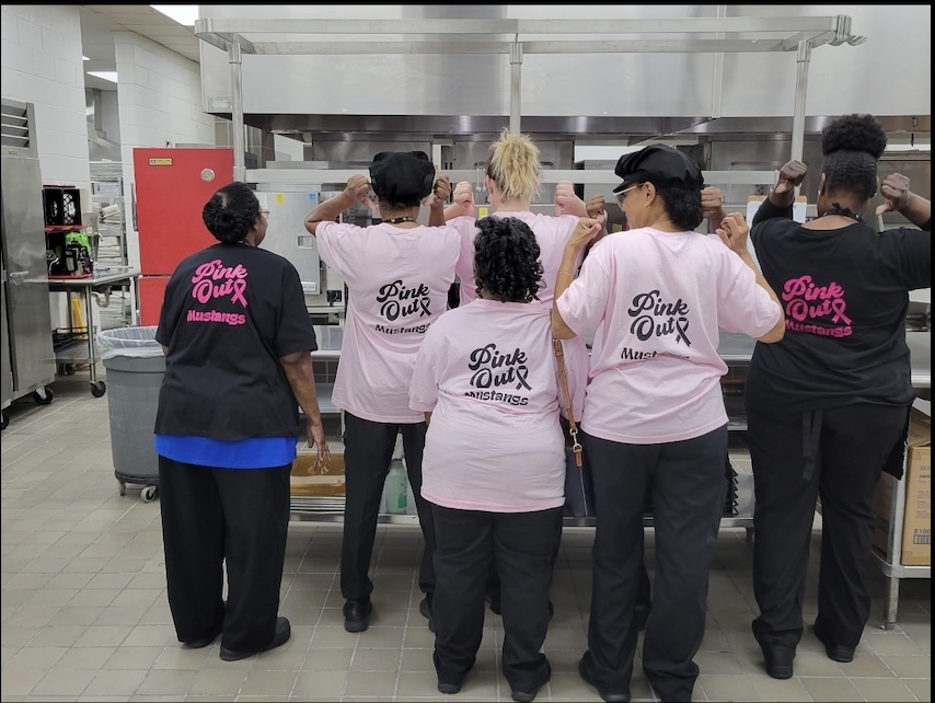A group of ladies wearing pink shirts and black pants facing backwards in a cafeteria.