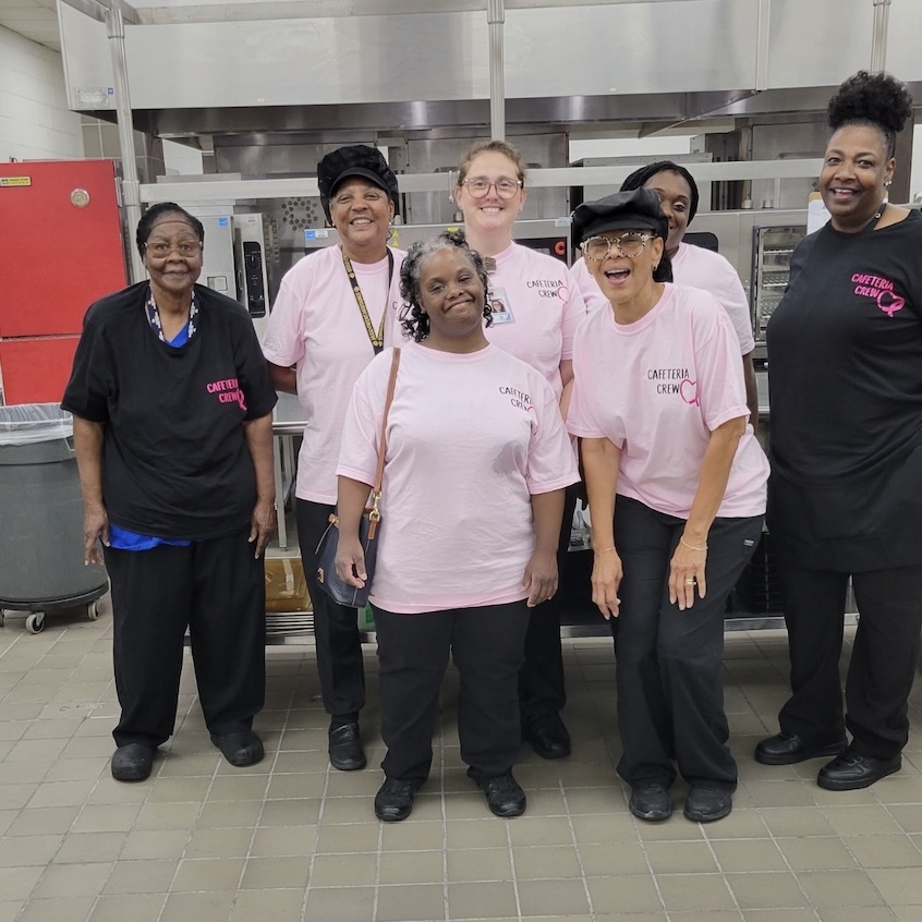 A group of ladies standing in a cafeteria wearing pink shirts and black pants.