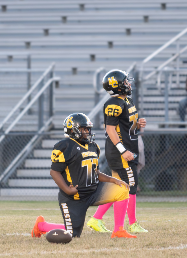 Two football players standing on a football field.  One is standing and one is kneeling. They are both wearing black and gold uniforms with pink socks.