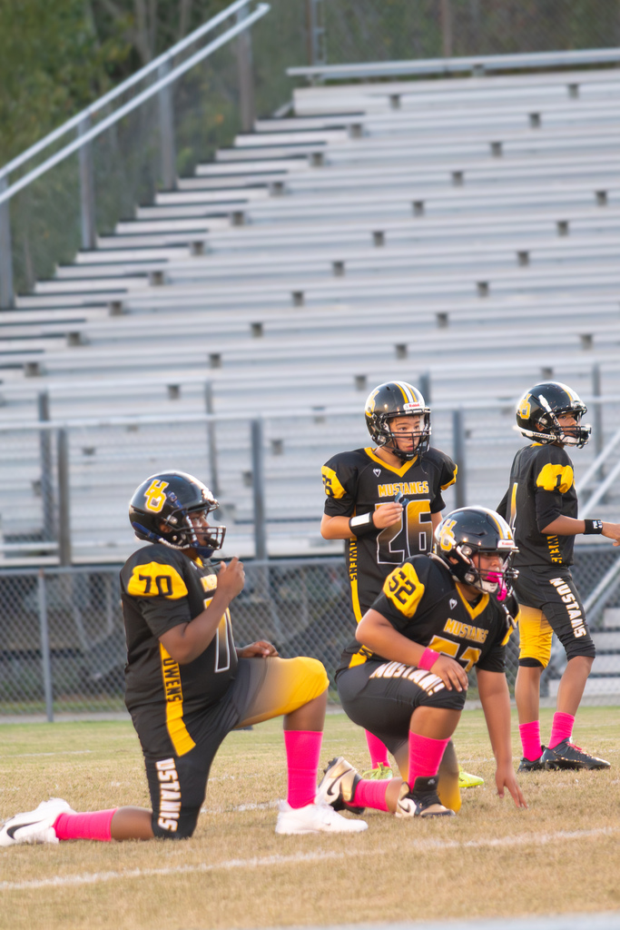 A close up picture of 4 football players on a field wearing black and gold uniforms with black helmets and pink socks.    Two of them are kneeling and 2 are standing,
