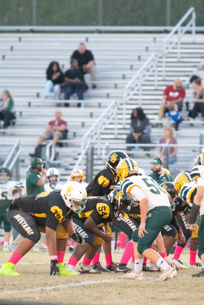 A close up picture of 2 football teams lined up to begin a play.   One group is wearing black and gold uniforms with black helmets.  The second team is wearing white and green uniforms