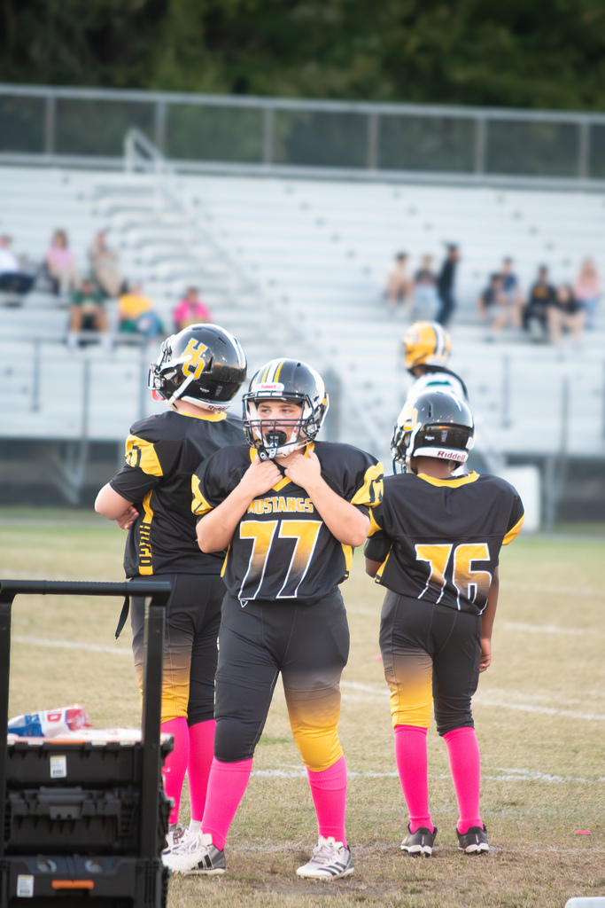 Close up picture of 3 football players outside on a football field wearing black and gold uniforms with pink socks and black helmets