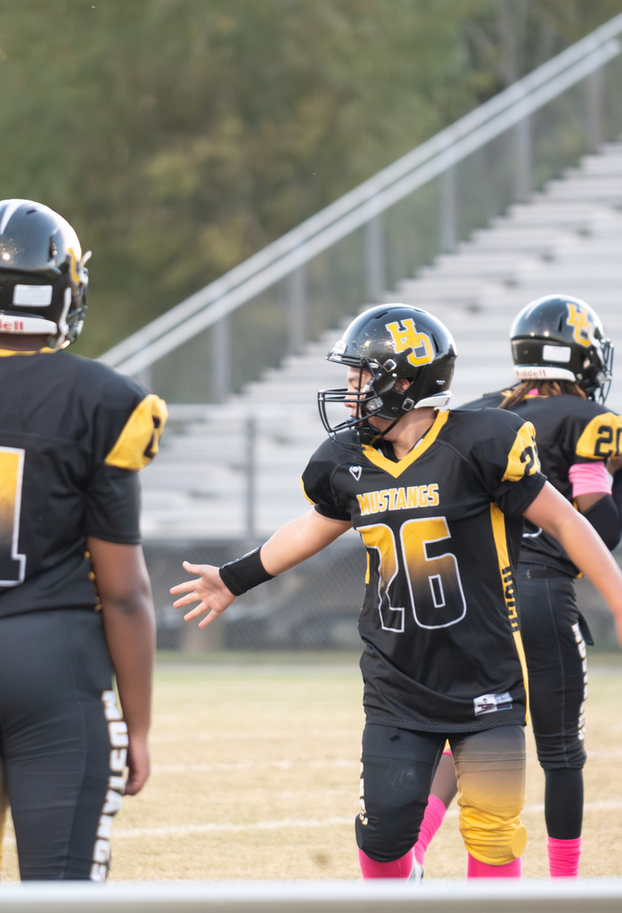 Three football payers standing outside on a football field. One is standing looking back with his arm extended.  They are wearing black and gold uniforms with black helmets.
