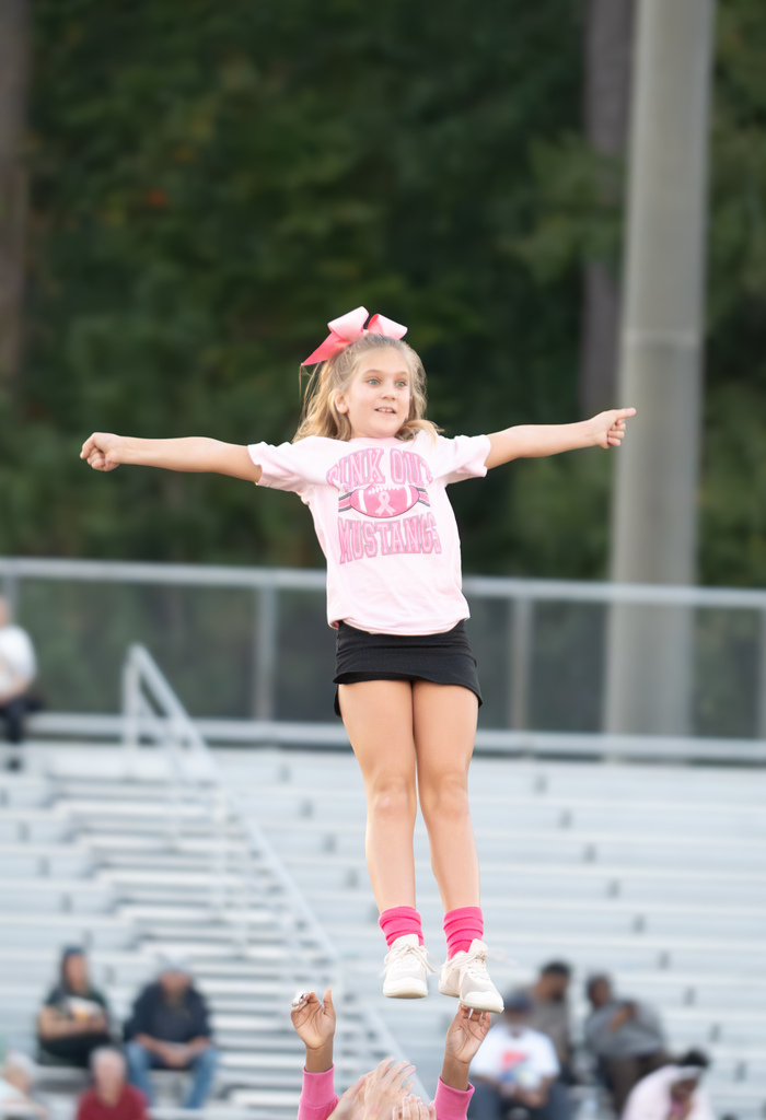 A cheerleader is being tossed in the air holding her arms out.  She is wearing a black skirt and a pink top.