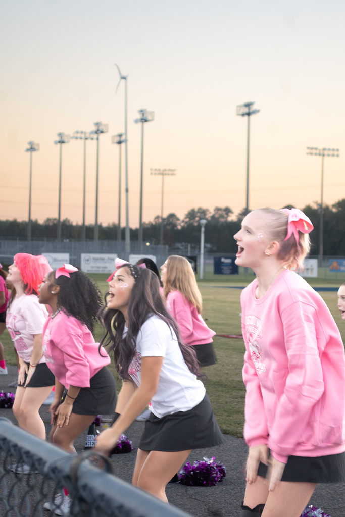 A group of cheerleaders are standing behind a fence with their legs slightly bent and hands clapping their thighs.  They are all wearing black skirts and pink shirts.
