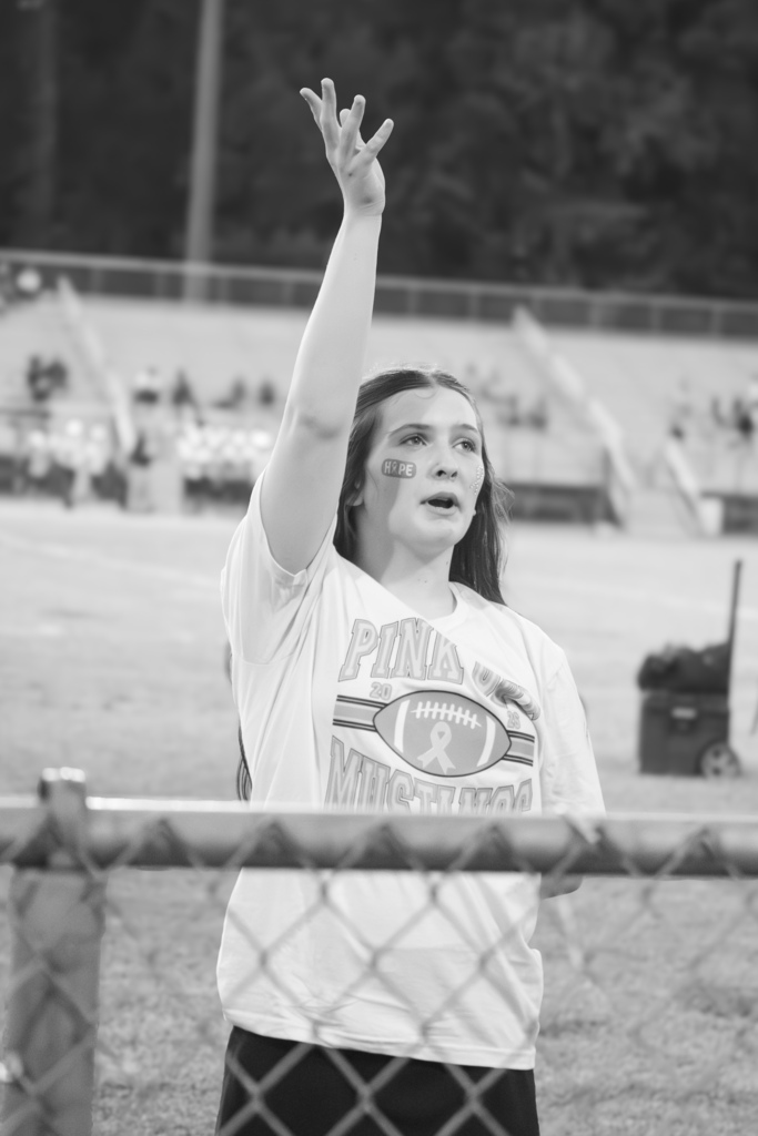 A cheerleader is standing behind a fence with one arm up in the air.  This is a black and white picture.