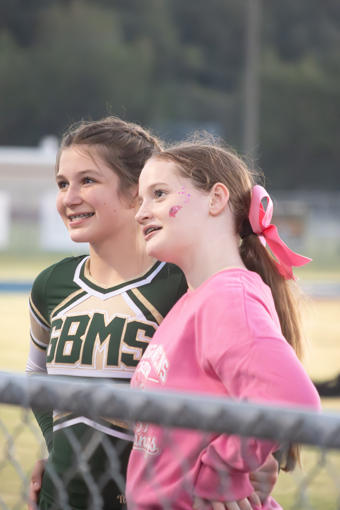 Two cheerleaders are standing behind a fence next to each other.  One is wearing a pink top and pink bow.  The other is wearing a green and gold uniform with GBMS letters on the top.